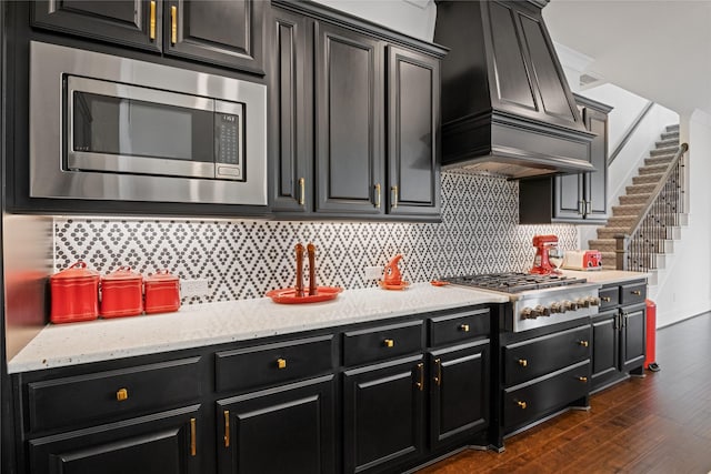 kitchen featuring dark cabinets, dark wood-type flooring, custom range hood, appliances with stainless steel finishes, and backsplash