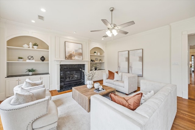 living room with built in features, a fireplace, ceiling fan, light hardwood / wood-style floors, and crown molding