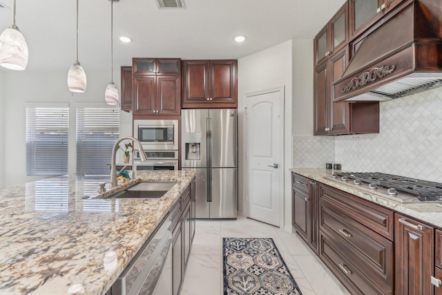 kitchen featuring appliances with stainless steel finishes, light stone counters, custom range hood, decorative light fixtures, and sink