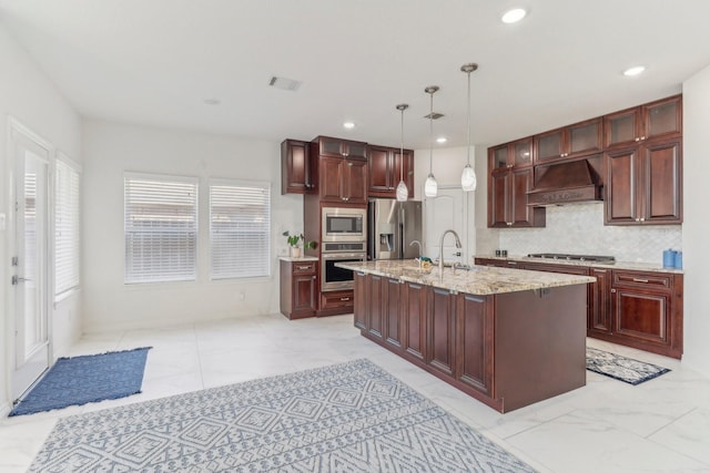 kitchen featuring an island with sink, stainless steel appliances, light stone countertops, decorative light fixtures, and custom exhaust hood