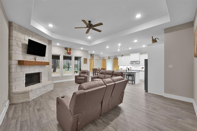 living room featuring ceiling fan, a tray ceiling, a fireplace, and light wood-type flooring