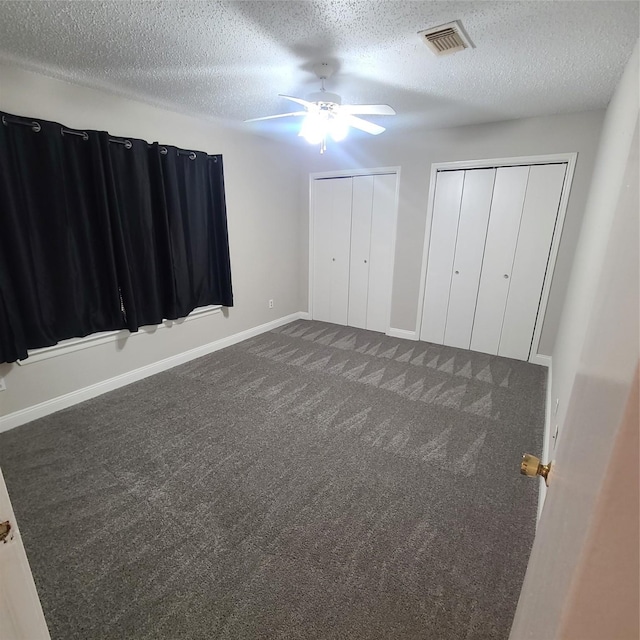 unfurnished bedroom featuring ceiling fan, dark carpet, a textured ceiling, and two closets