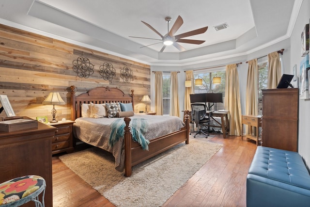 bedroom with hardwood / wood-style flooring, wooden walls, ceiling fan, a raised ceiling, and visible vents