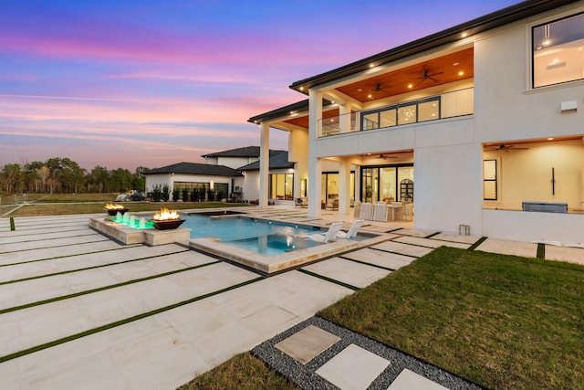 pool at dusk featuring a fire pit, a lawn, a patio, ceiling fan, and an infinity pool