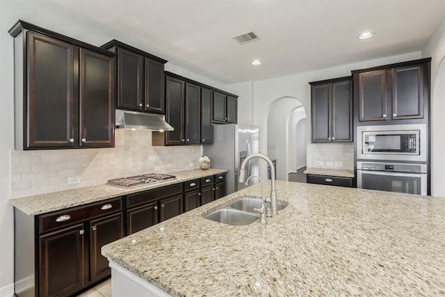 kitchen with sink, appliances with stainless steel finishes, light stone counters, dark brown cabinetry, and decorative backsplash