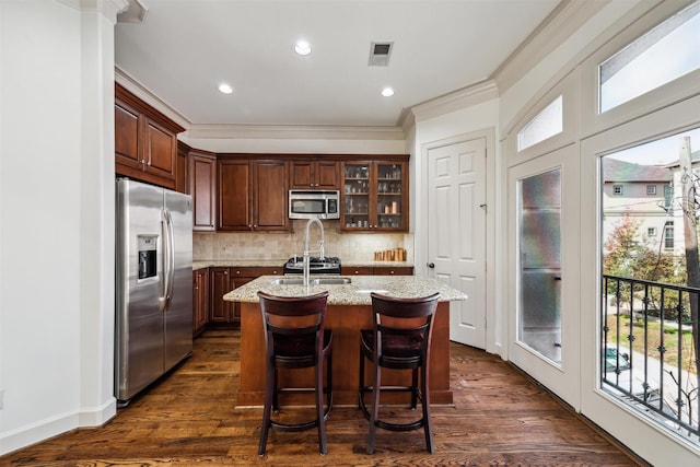 kitchen with ornamental molding, stainless steel appliances, light stone countertops, a kitchen island with sink, and decorative backsplash