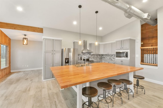 kitchen with an island with sink, stainless steel appliances, gray cabinets, wall chimney exhaust hood, and pendant lighting