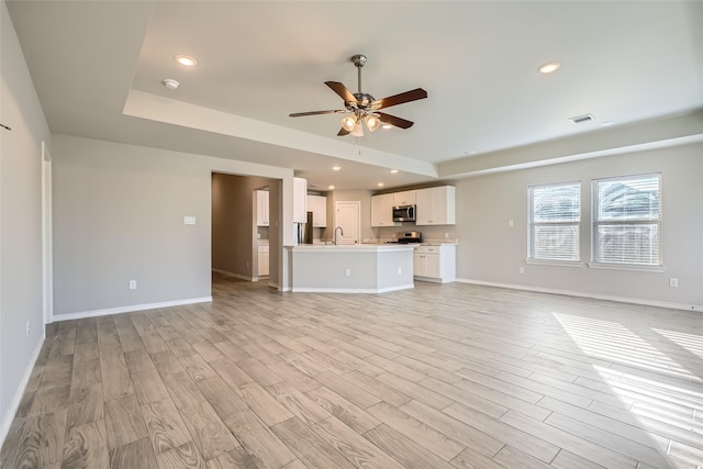 unfurnished living room with ceiling fan, a tray ceiling, sink, and light wood-type flooring