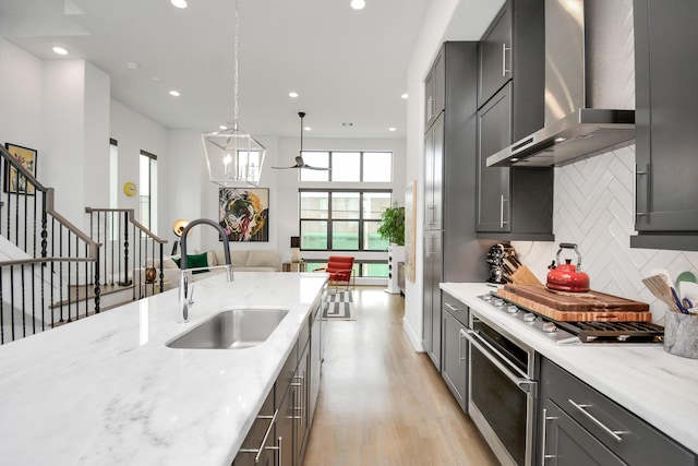 kitchen with stainless steel appliances, light stone countertops, sink, wall chimney range hood, and pendant lighting