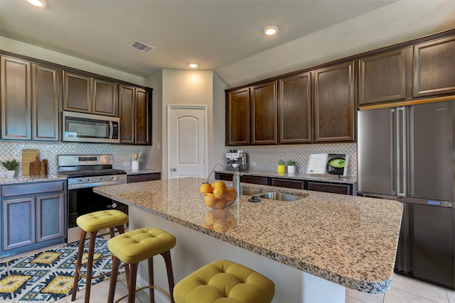 kitchen with a breakfast bar area, dark brown cabinetry, stainless steel appliances, light stone countertops, and a center island with sink