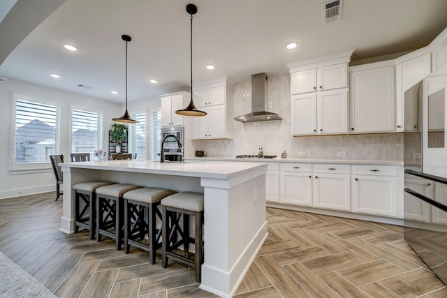 kitchen with pendant lighting, white cabinetry, an island with sink, and wall chimney range hood