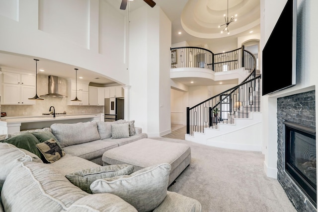 living room with a stone fireplace, ceiling fan with notable chandelier, sink, light colored carpet, and a tray ceiling