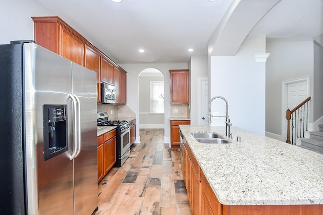 kitchen with sink, an island with sink, stainless steel appliances, light stone countertops, and light hardwood / wood-style floors