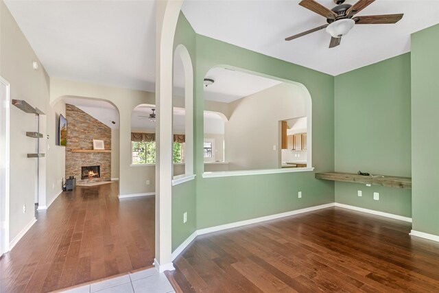unfurnished room featuring hardwood / wood-style floors, ceiling fan, a stone fireplace, and arched walkways