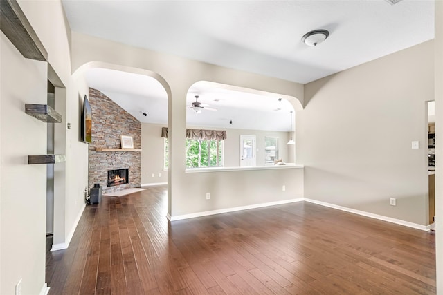 unfurnished living room featuring baseboards, arched walkways, dark wood-style flooring, ceiling fan, and a stone fireplace