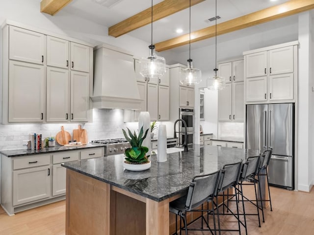 kitchen with decorative light fixtures, dark stone counters, an island with sink, custom range hood, and stainless steel appliances