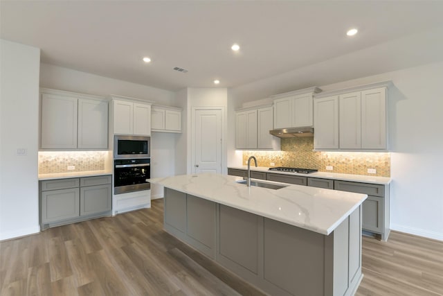 16119 Rustling Woods Rd, Conroe, TX 77302 - kitchen featuring stainless steel appliances, tasteful backsplash, light stone countertops, light wood-style flooring, and a center island with sink
