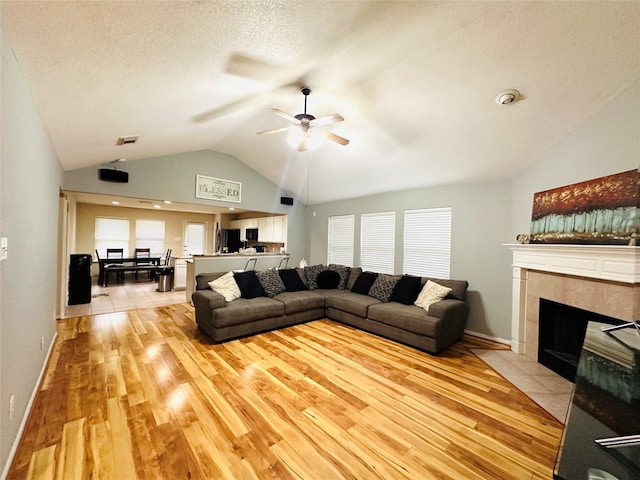 living room with ceiling fan, light hardwood / wood-style floors, a textured ceiling, a tiled fireplace, and vaulted ceiling