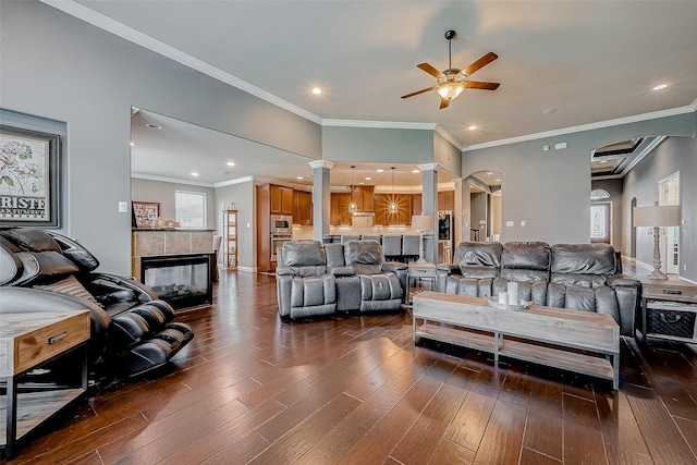 living room featuring ornate columns, dark hardwood / wood-style floors, a tiled fireplace, ceiling fan, and crown molding