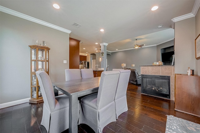 dining space with crown molding, ceiling fan, dark hardwood / wood-style floors, a tiled fireplace, and ornate columns