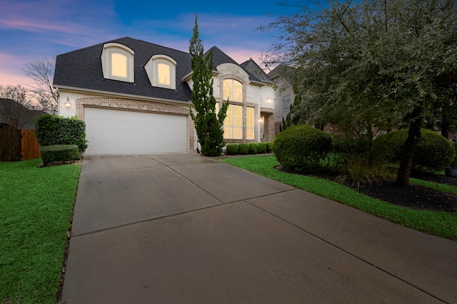 french country inspired facade with a garage, a shingled roof, brick siding, driveway, and a yard