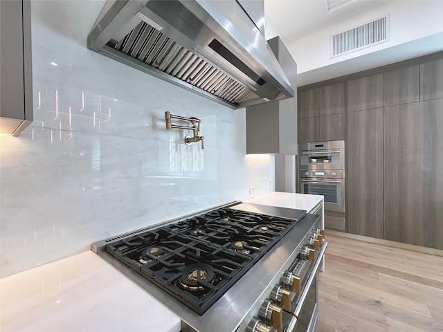 kitchen featuring wall chimney exhaust hood, cooktop, stainless steel double oven, light hardwood / wood-style floors, and decorative backsplash