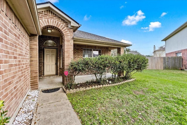 property entrance featuring roof with shingles, brick siding, a lawn, and fence