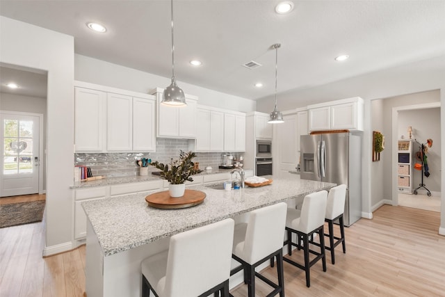 kitchen featuring an island with sink, hanging light fixtures, stainless steel appliances, backsplash, and white cabinets