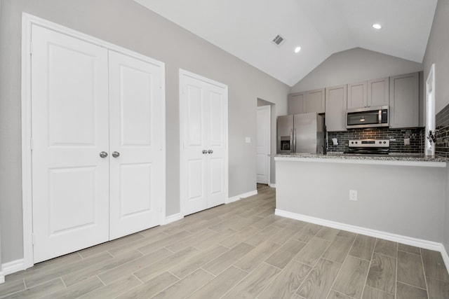 kitchen featuring gray cabinets, stainless steel appliances, tasteful backsplash, light stone countertops, and vaulted ceiling