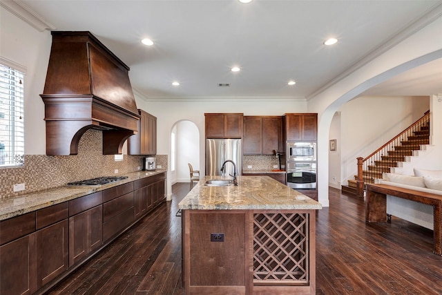 kitchen featuring arched walkways, a kitchen island with sink, a sink, appliances with stainless steel finishes, and light stone countertops