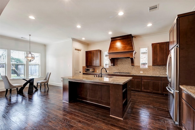 kitchen featuring visible vents, an island with sink, premium range hood, pendant lighting, and a sink