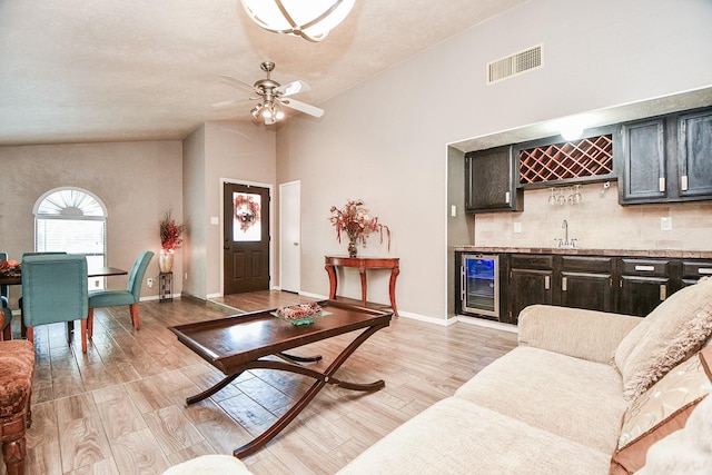 living room with indoor wet bar, ceiling fan, vaulted ceiling, beverage cooler, and light wood-type flooring