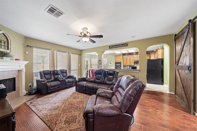 living room featuring a tiled fireplace, light hardwood / wood-style flooring, a barn door, and ceiling fan