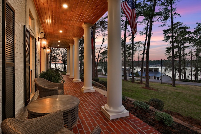 patio terrace at dusk featuring a lawn, a water view, and covered porch