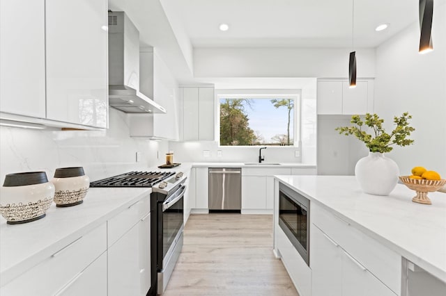 kitchen featuring wall chimney range hood, sink, hanging light fixtures, stainless steel appliances, and white cabinets