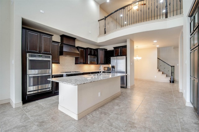 kitchen featuring premium range hood, sink, appliances with stainless steel finishes, an island with sink, and light stone countertops