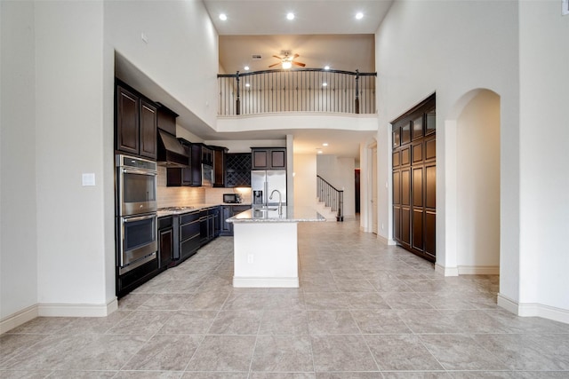 kitchen featuring dark brown cabinets, stainless steel appliances, light stone counters, custom range hood, and an island with sink