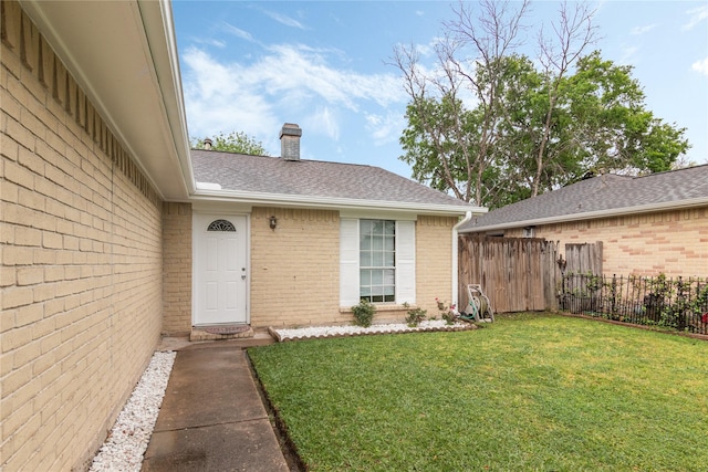 property entrance with roof with shingles, a chimney, fence, brick siding, and a lawn