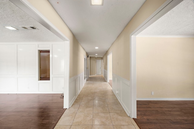 hallway with a textured ceiling, light wood-style flooring, visible vents, and a decorative wall