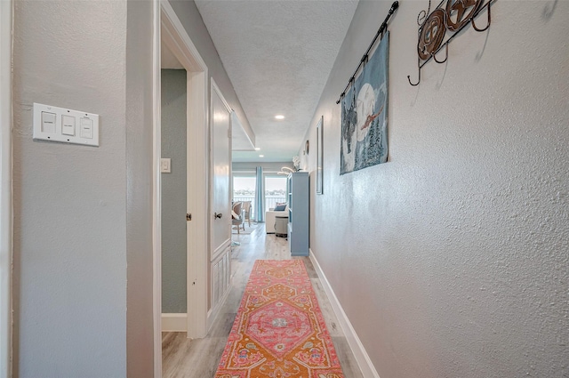 hallway with baseboards, a textured wall, a textured ceiling, and light wood-style floors