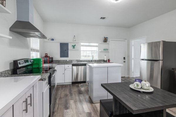 kitchen with appliances with stainless steel finishes, white cabinetry, a center island, electric panel, and wall chimney range hood