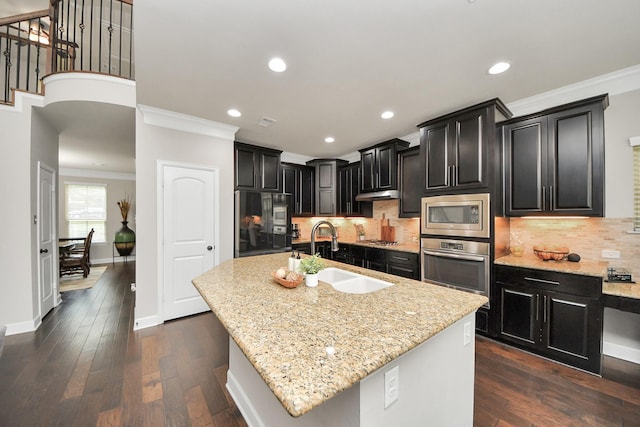 kitchen with sink, a kitchen island with sink, built in appliances, light stone counters, and dark hardwood / wood-style flooring