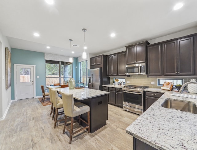 kitchen with dark brown cabinetry, appliances with stainless steel finishes, light stone counters, hanging light fixtures, and a sink