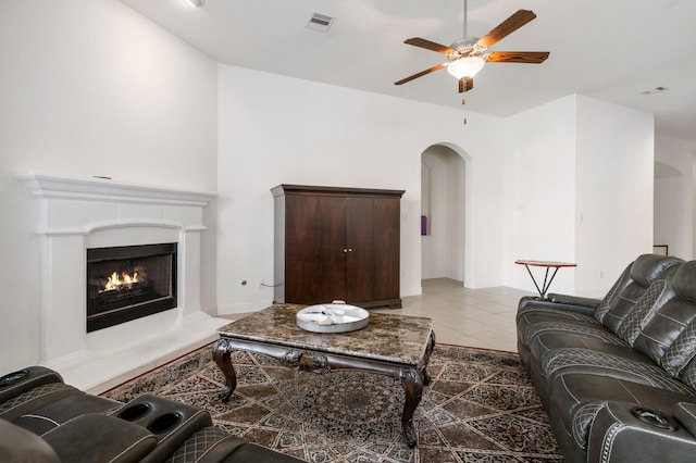 living room with light tile patterned floors, arched walkways, a ceiling fan, a warm lit fireplace, and visible vents