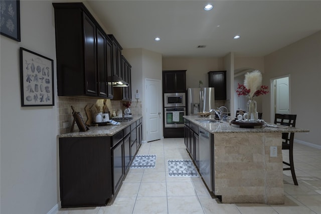 kitchen with an island with sink, sink, backsplash, light stone counters, and stainless steel appliances