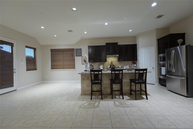 kitchen featuring appliances with stainless steel finishes, a kitchen island with sink, tasteful backsplash, light stone countertops, and a kitchen bar