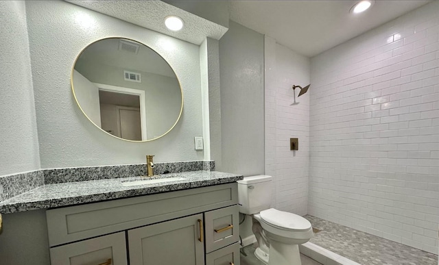 bathroom with visible vents, toilet, a tile shower, a textured wall, and vanity