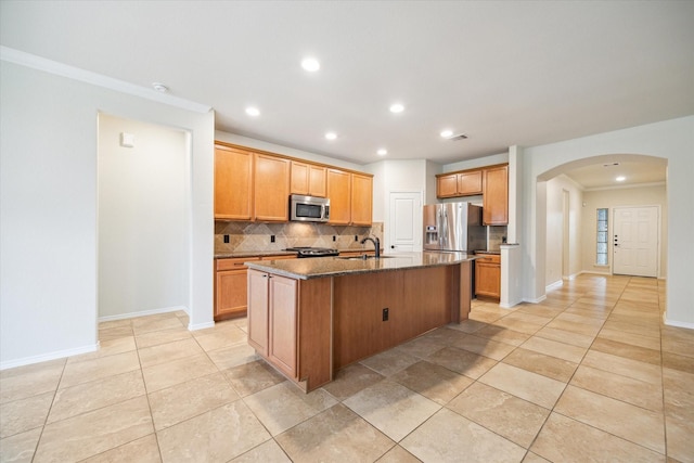 kitchen with a sink, decorative backsplash, arched walkways, stainless steel appliances, and a kitchen island with sink