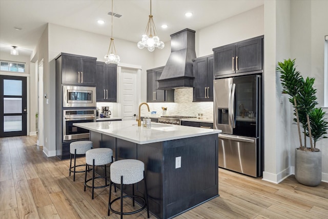 kitchen featuring light wood-type flooring, a center island with sink, hanging light fixtures, stainless steel appliances, and premium range hood