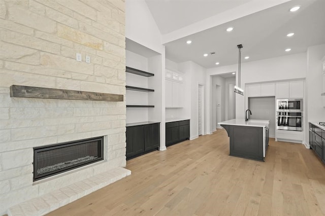 kitchen featuring built in microwave, a stone fireplace, an island with sink, stainless steel oven, and white cabinetry